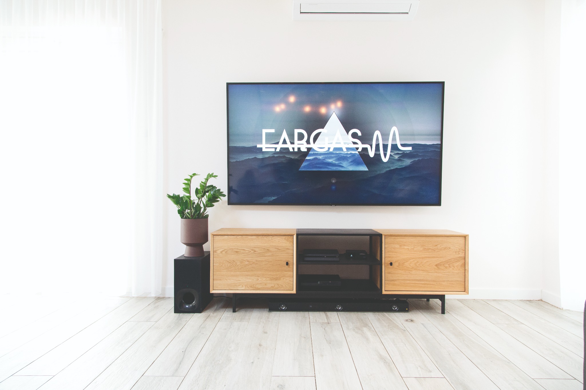 Nowoczesny salon z telewizorem wyświetlającym abstrakcyjny obraz, drewnianą szafką RTV na czarnych nóżkach, subwooferem i rośliną doniczkową na jasnej podłodze.