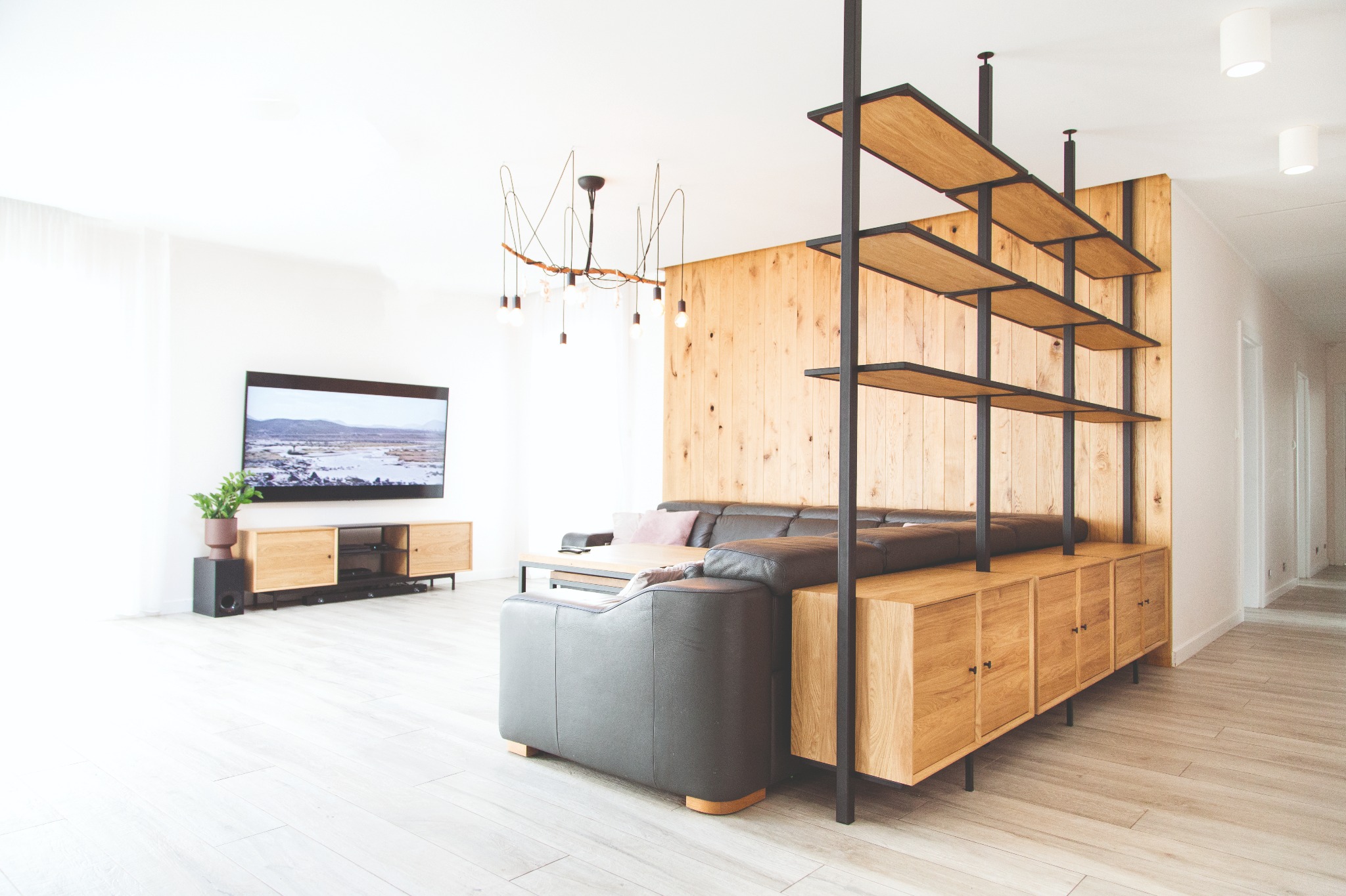 Nowoczesny salon z drewnianą ścianą, regałem z metalowymi elementami i drewnianymi półkami oraz szarą sofą.