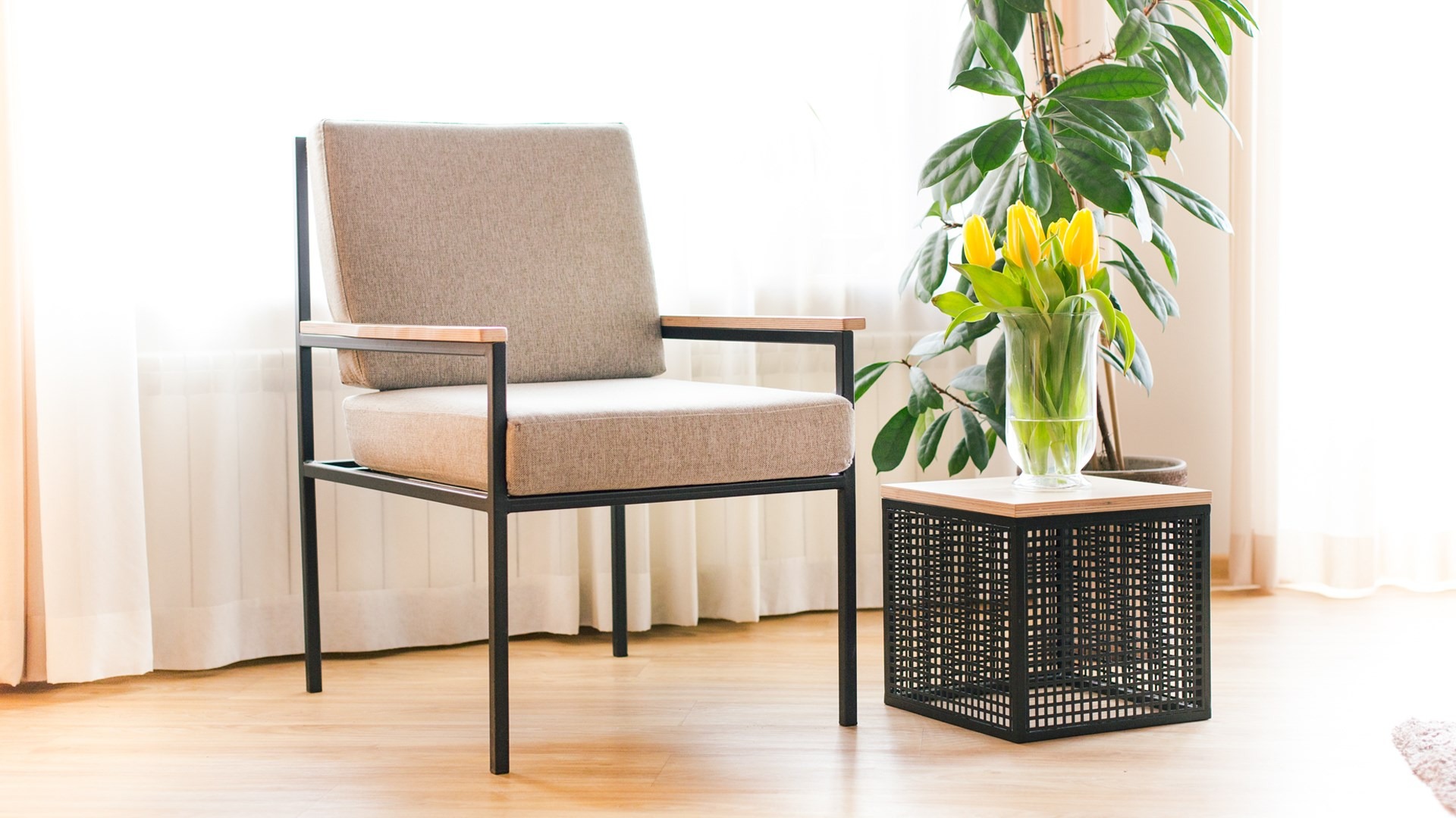 Nowoczesne krzesło z metalową ramą i tapicerowanym siedziskiem obok ażurowego stolika z tulipanami w szklanym wazonie, na jasnym drewnianym parkiecie przy oknie z białymi zasłonami.