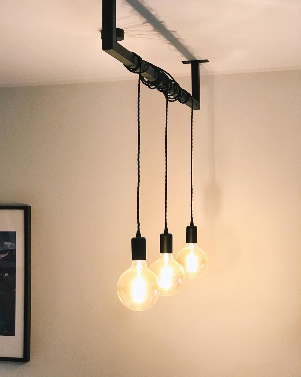 Minimalistyczna lampa sufitowa z trzema żarówkami Edisona zawieszonymi na czarnych kablach, przymocowanych do metalowej belki, oświetlona ciepłym światłem, widoczna na tle jasnej ściany.