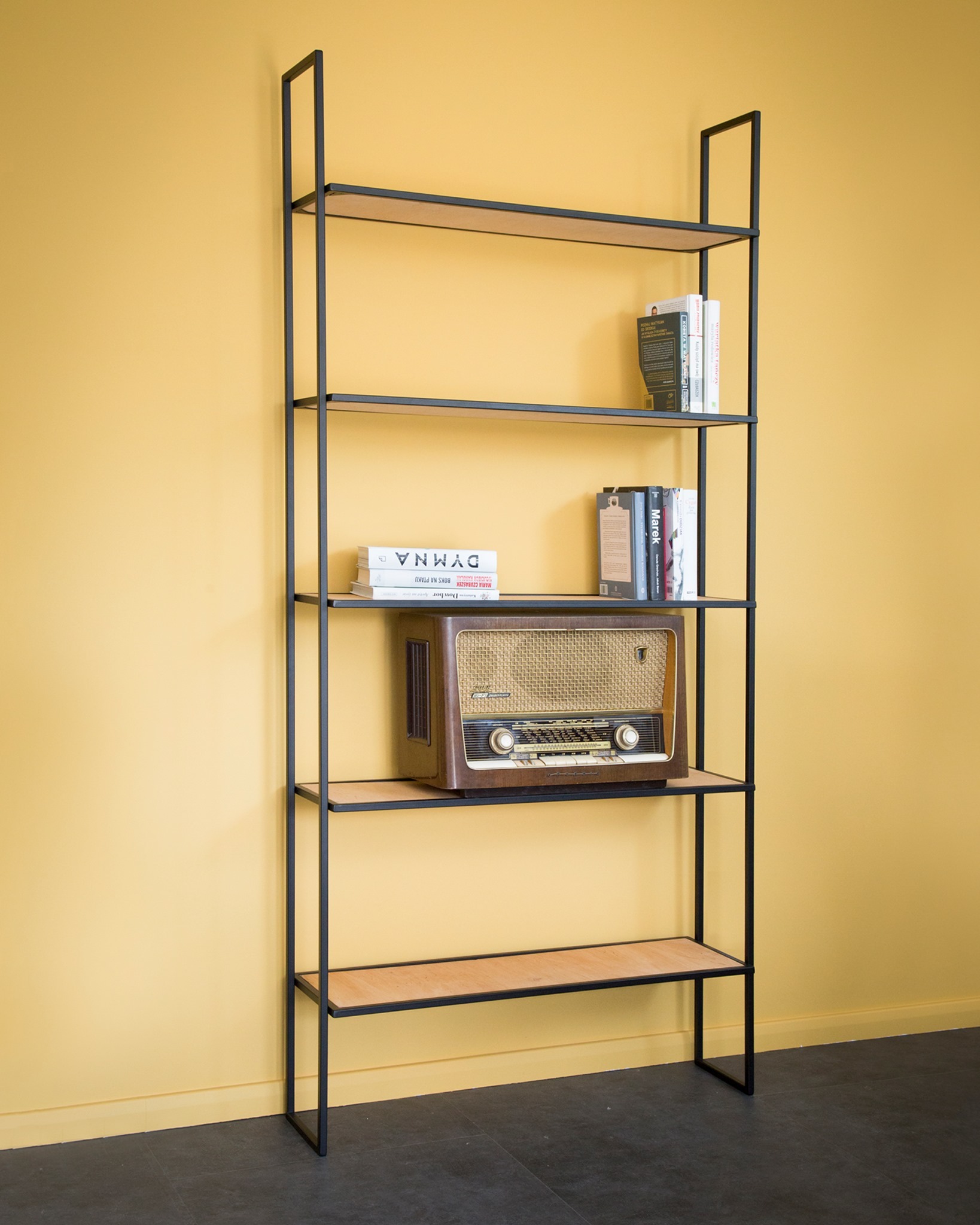 Wysoki, nowoczesny regał z czarną metalową ramą i jasnymi drewnianymi półkami, na których stoją książki i zabytkowe radio, na tle żółtej ściany.