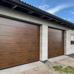 Dwie brązowe bramy garażowe segmentowe z poziomym wzorem drewna, zamontowane w surowym budynku z bloczków betonowych, widok z zewnątrz.