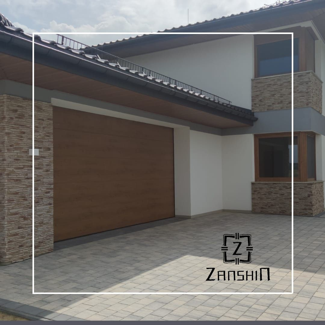 Nowoczesny dom z brązową bramą garażową segmentową i elewacją z kamienia dekoracyjnego. Brama z poziomymi panelami. Jasna kostka brukowa na podjeździe.