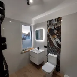 Nowoczesna łazienka z drewnopodobną podłogą, białymi płytkami ściennymi, umywalką z szafką, toaletą i dekoracyjną pionową płytką imitującą marmur. Nad umywalką podświetlane lustro, a nad oknem małe...