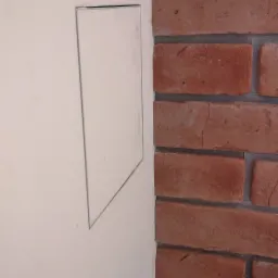 Biała ściana z ukrytymi drzwiczkami rewizyjnymi sąsiadująca z dekoracyjną ścianą z czerwonej cegły.