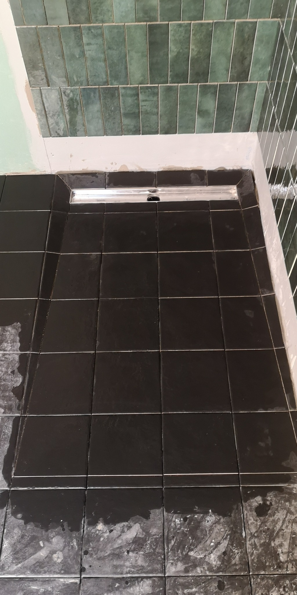 Ułożona, czarna, kwadratowa podłoga z płytek w kabinie prysznicowej z odpływem liniowym; zielone, pionowe płytki na ścianie w tle, widoczne ślady po fugowaniu.