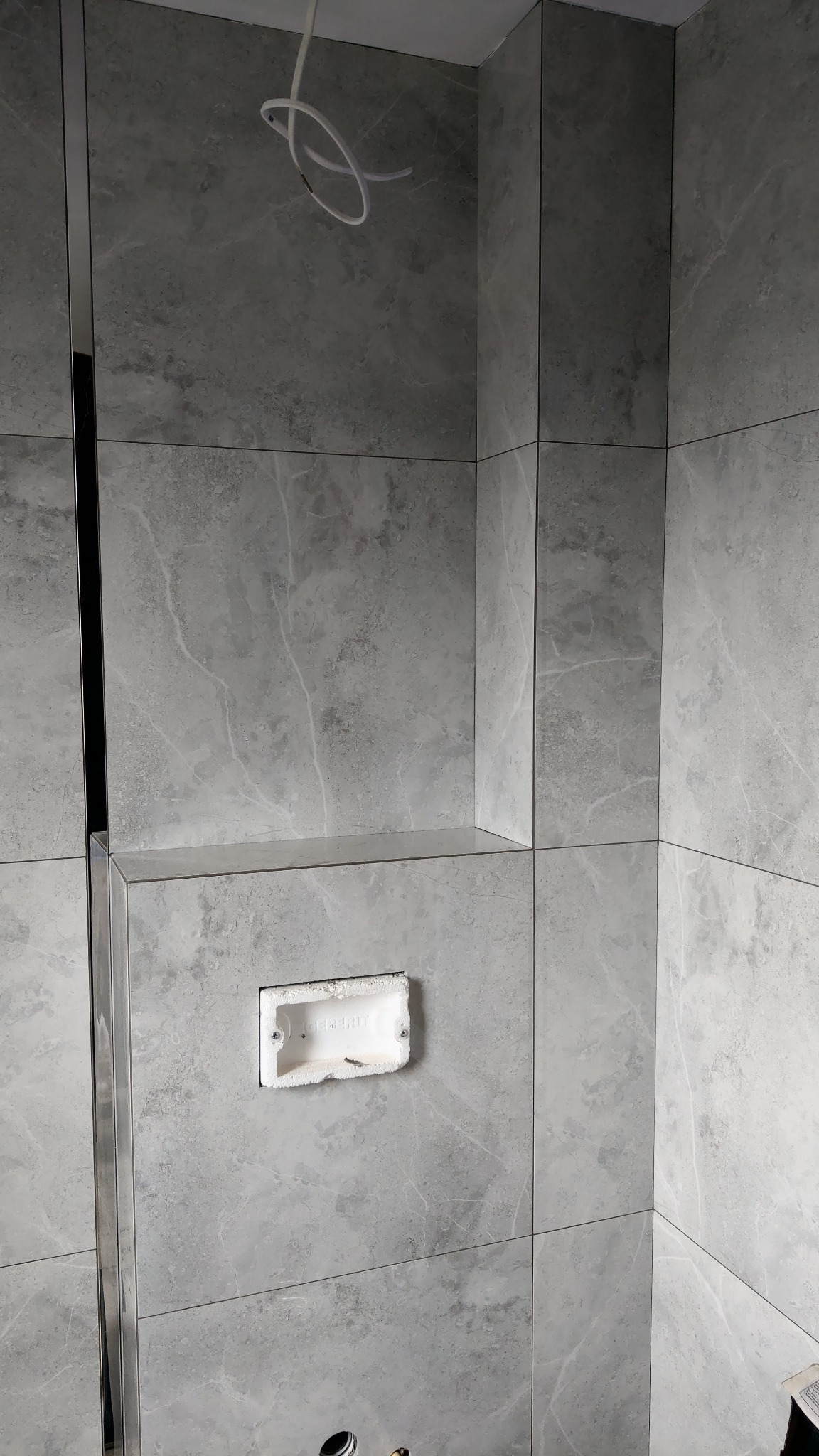 Ściana łazienki wyłożona szarymi płytkami imitującymi marmur, z widoczną puszką instalacyjną podtynkową, chromowana listwa dekoracyjna pionowa i wystające kable z sufitu.