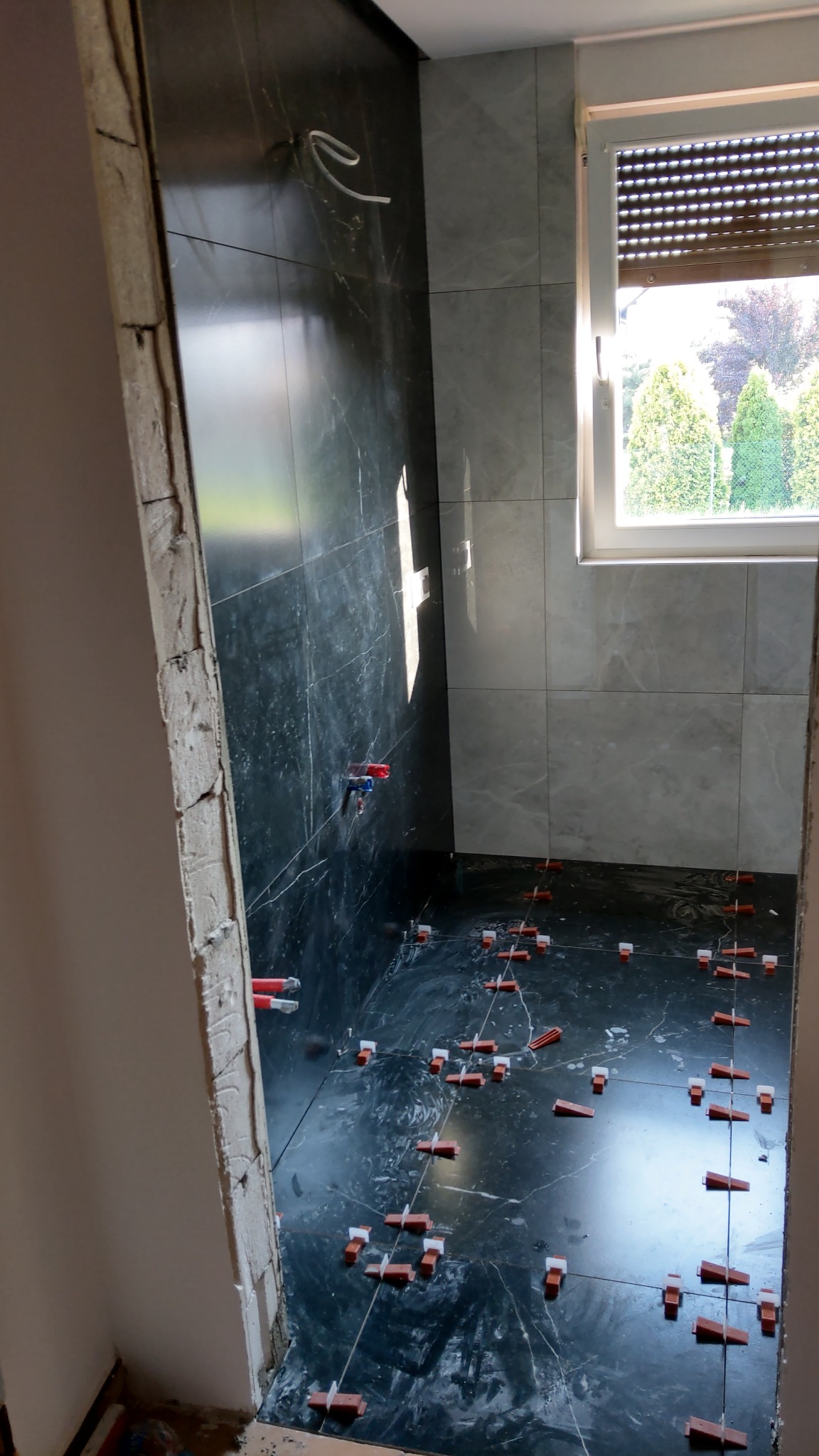 Wnętrze łazienki w trakcie układania ciemnych płytek z marmurowym wzorem na podłodze i ścianach, widoczne krzyżyki dystansowe, surowe wykończenie ościeżnicy drzwiowej, instalacja wodna w ścianie...