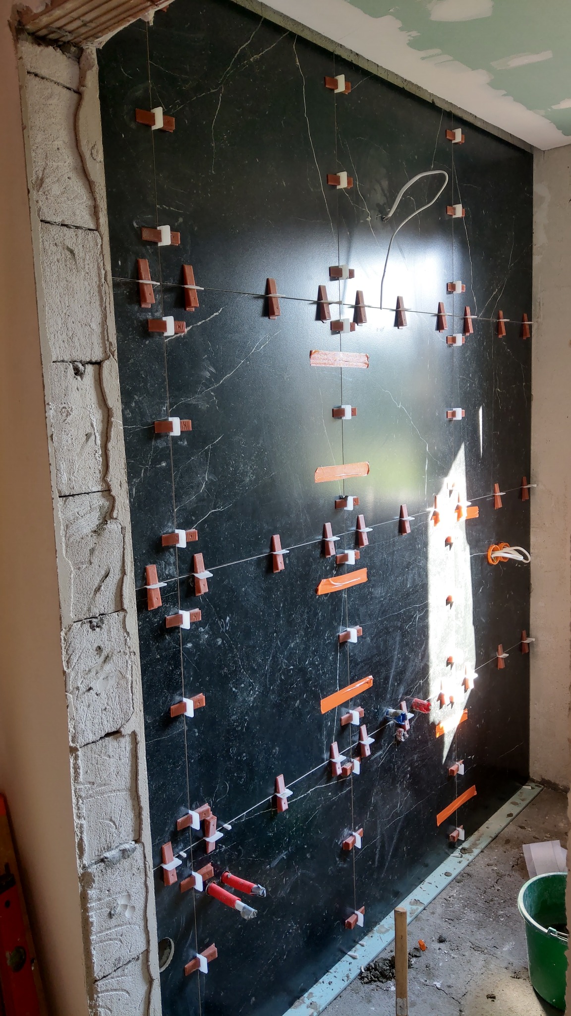 Ściana z czarnych płyt marmuropodobnych w trakcie montażu, z widocznymi systemami poziomowania płytek: klipsami, klinami i sznurkami, oraz fragmentem surowej ściany po lewej stronie.