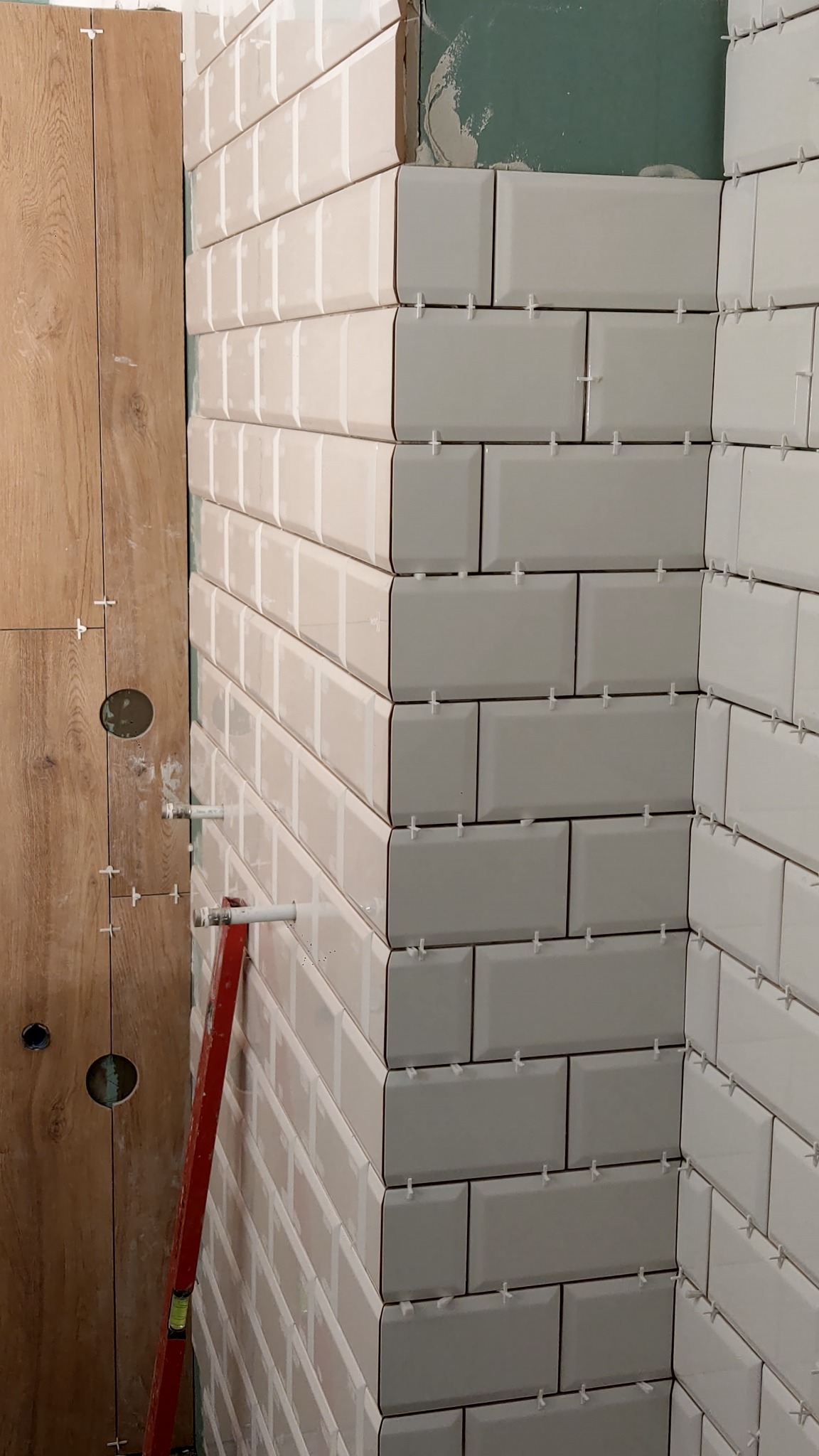 Białe, prostokątne płytki ułożone na ścianie z krzyżykami dystansowymi, widoczny fragment płyty kartonowo-gipsowej i poziomica oparta o ścianę z drewnopodobnej płyty.