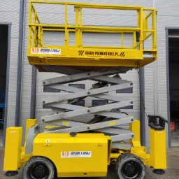 Żółty podnośnik nożycowy marki Amar i Maj, z platformą roboczą, zaparkowany na kostce brukowej przed szarą halą z bramą rolowaną.