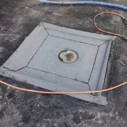 Szczegółowe ujęcie odpływu dachowego zabezpieczonego papą, z widocznymi łączeniami i elementami uszczelniającymi, otoczonego bitumiczną powierzchnią dachu i fragmentem węża.