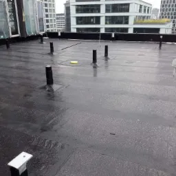 Płaski dach pokryty czarną membraną bitumiczną, widoczne pionowe rury i elementy mocujące, w tle nowoczesne budynki biurowe.
