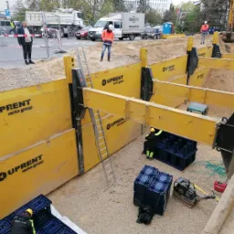 Głęboki wykop zabezpieczony żółtymi szalunkami UPRENT, pracownicy w odblaskowych ubraniach, ubijarka BOMAG i stosy plastikowych skrzynek na żwirowym dnie wykopu, na górze widoczny ruch uliczny...