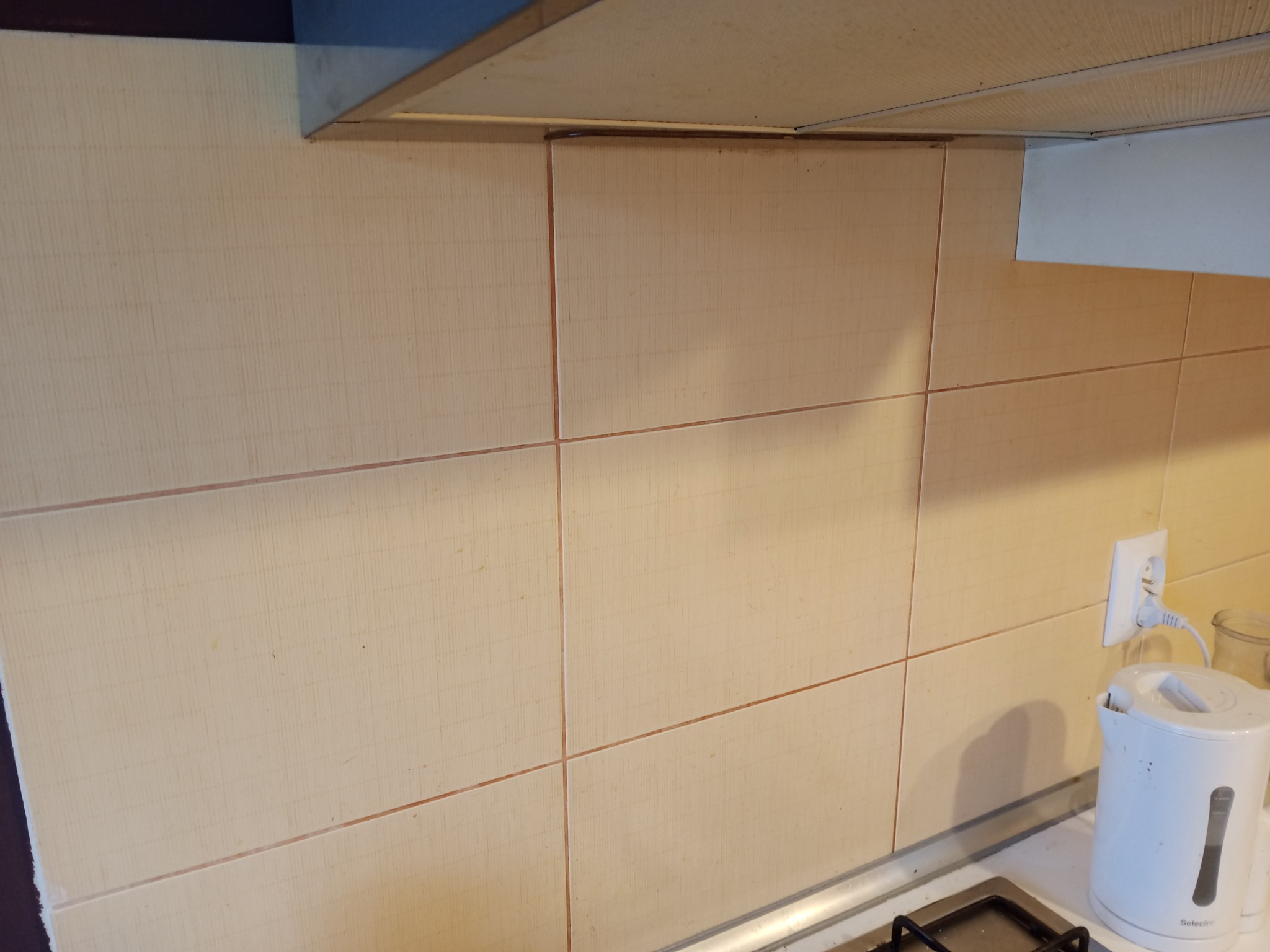 Beżowe płytki ścienne z delikatnym wzorem pionowych prążków, ułożone w kuchni pod okapem, widoczny fragment płyty grzewczej i czajnik elektryczny.