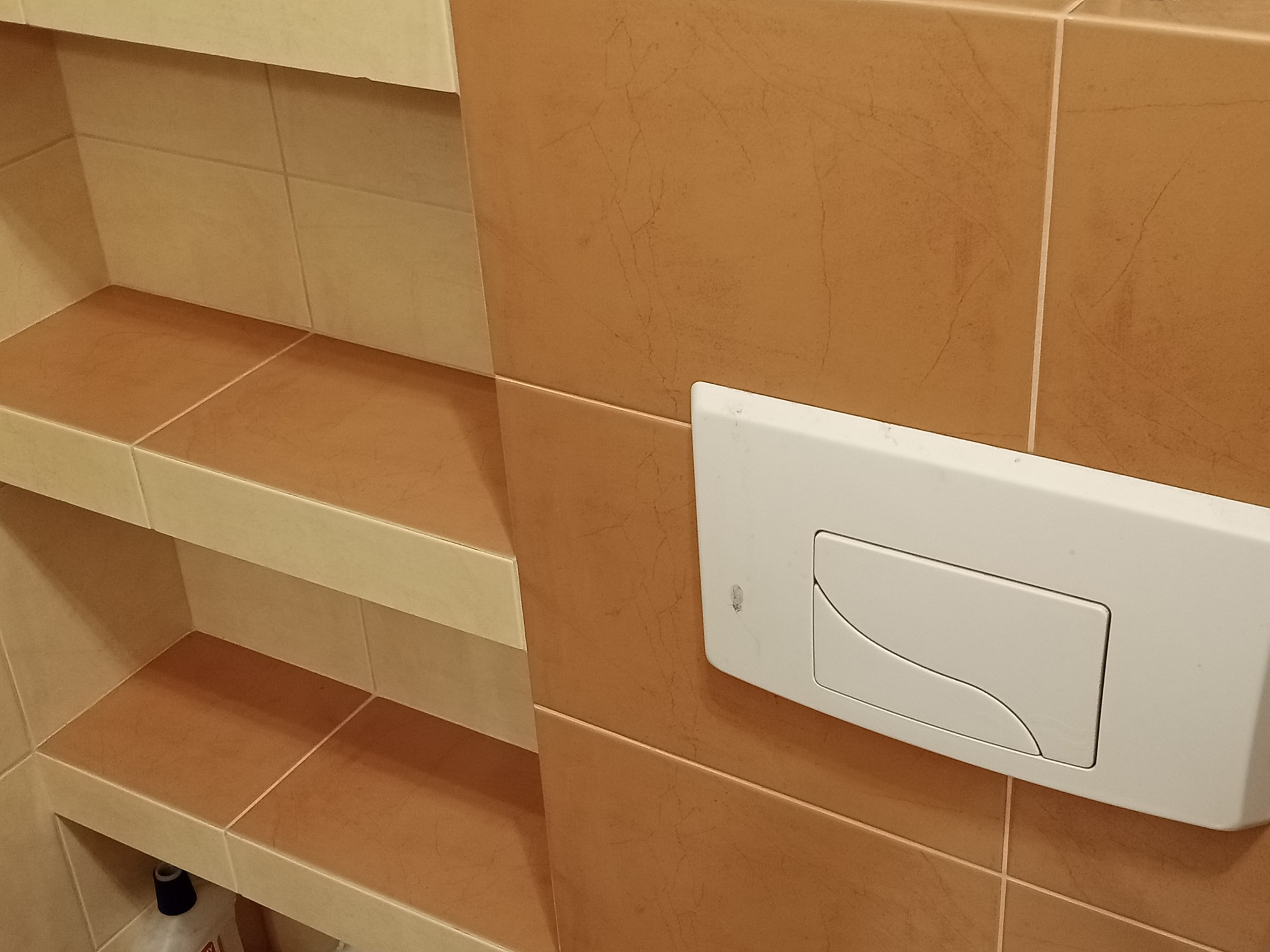 Beżowo-brązowe płytki ceramiczne w łazience z wnęką półkową i białym przyciskiem spłuczki podtynkowej na ścianie.