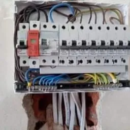 Otwarta skrzynka rozdzielcza z zamontowanymi bezpiecznikami i przewodami o różnych kolorach, wystająca ze ściany z widocznym fragmentem cegły. Przewody uporządkowane wewnątrz skrzynki, ale...