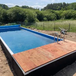 Prostokątny basen naziemny z drewnianym pomostem i metalową drabinką, widok na ogród i drzewa w tle, częściowo zakryty niebieską plandeką.