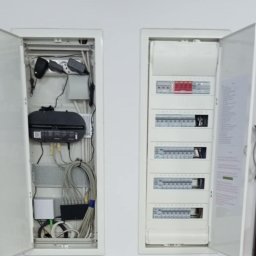 Rafał Skiba - Dwie otwarte, wbudowane w ścianę szafki: po lewej panel multimedialny z routerem i uporządkowanym okablowaniem sieciowym, po prawej rozdzielnica elektryczna z modułami zabezpieczającymi.