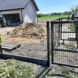 Marcin Kuklinski Montaż Ogrodzen - Czarny metalowy płot panelowy z otwartą furtką na tle budowy domu i zielonego krajobrazu, widoczny mężczyzna przy samochodzie dostawczym.