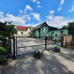 Czarna brama wjazdowa z siatki na posesję z budynkami w tle, dwa kwietniki z białymi kwiatami po bokach bramy, kostka brukowa na podjeździe, widok za dnia przy słonecznej pogodzie.