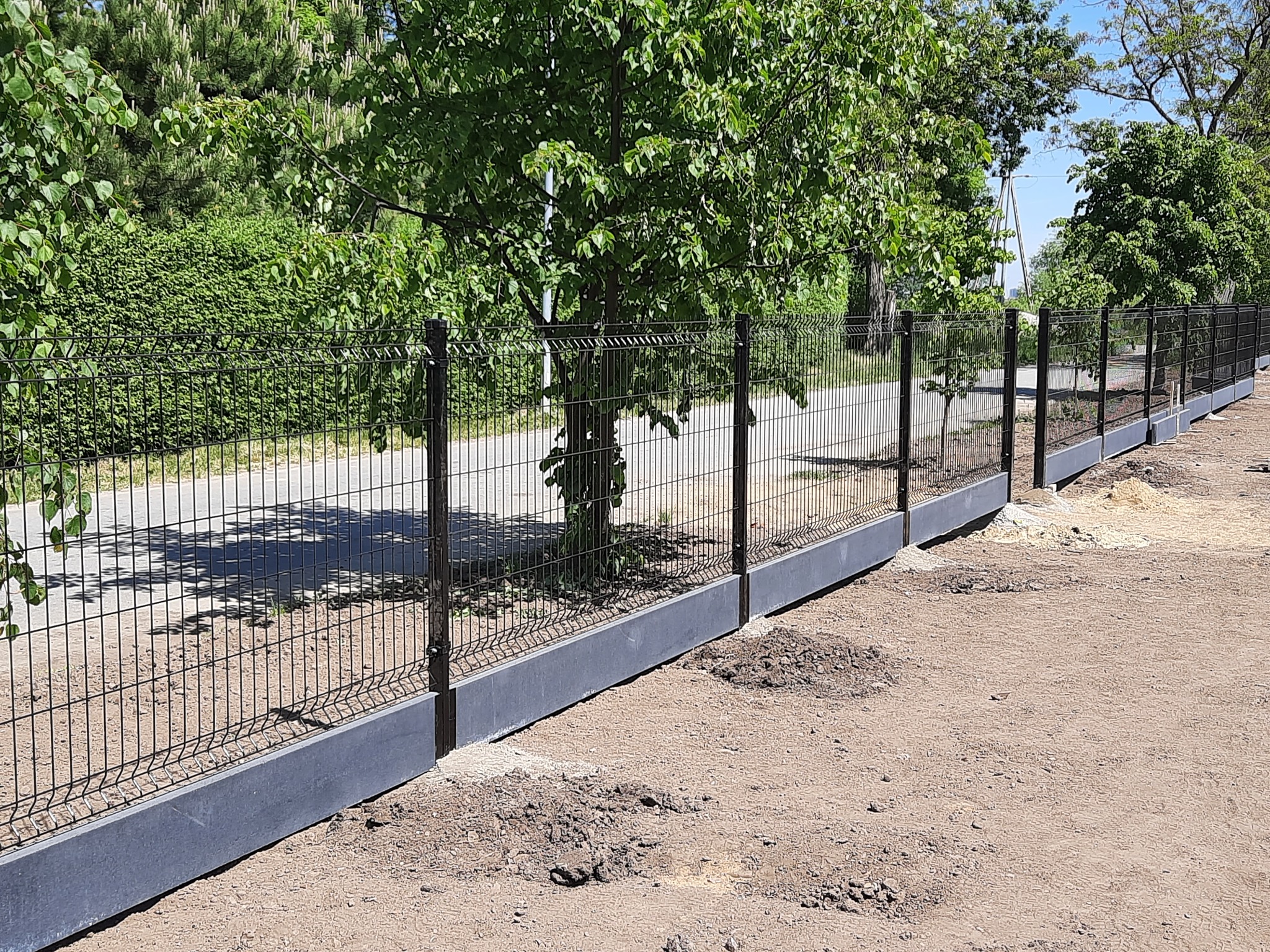 Długi, czarny metalowy płot panelowy z betonową podmurówką, biegnący wzdłuż drogi i zieleni, w słoneczny dzień.