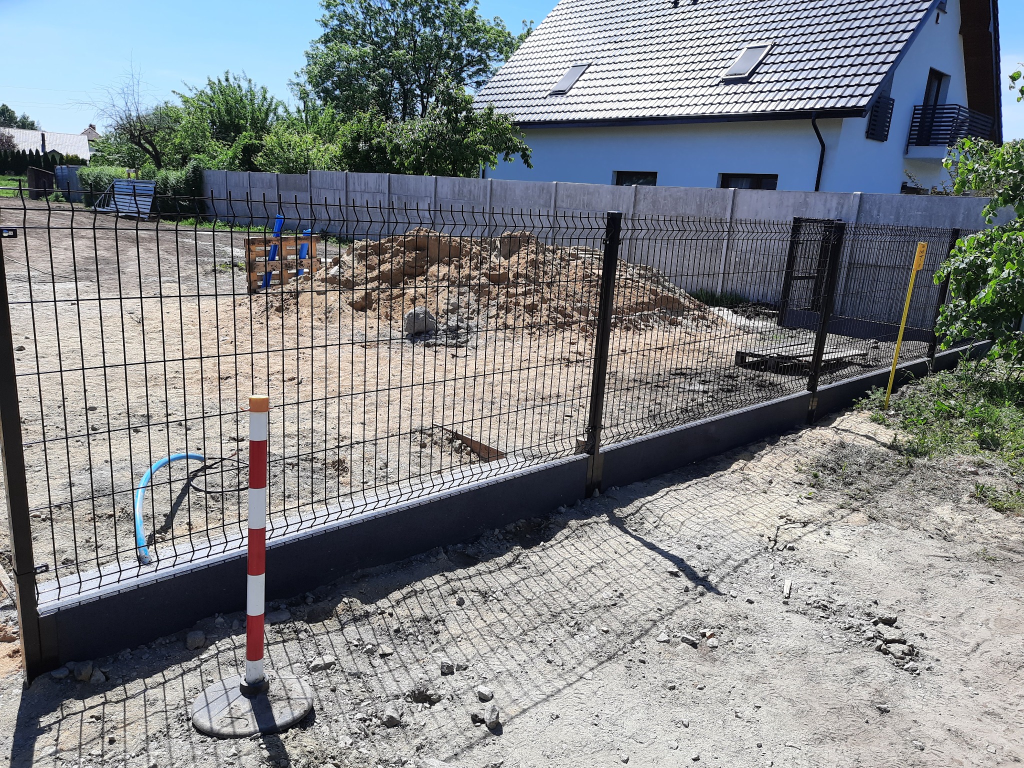 Czarny panelowy system ogrodzeniowy z podmurówką betonową w trakcie montażu, widoczny stos ziemi i narzędzia, w tle dom jednorodzinny.