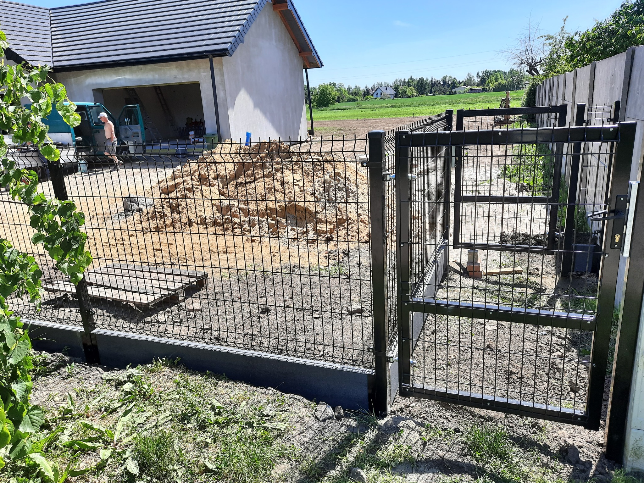 Czarny metalowy płot panelowy z otwartą furtką na tle budowy domu i zielonego krajobrazu, widoczny mężczyzna przy samochodzie dostawczym.