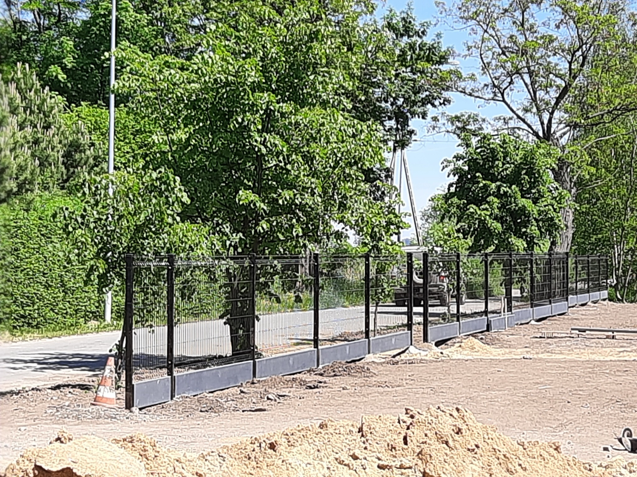 Nowo postawione, czarne, metalowe ogrodzenie z podmurówką na tle zieleni, z widocznym stożkiem ostrzegawczym i stertą piasku, sugerujące trwające prace wykończeniowe.