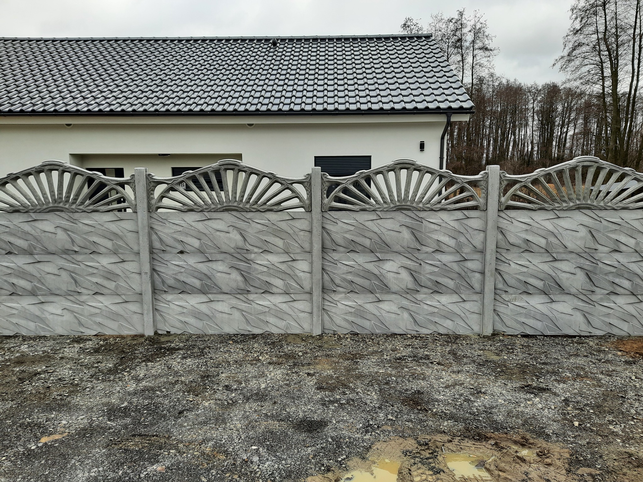 Dekoracyjny, szary, betonowy płot z motywem słońca w Wołowie, widoczny na tle białego domu z szarym dachem i drzew bez liści.