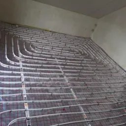 Rozłożona instalacja ogrzewania podłogowego z białych rur na folii z siatką w surowym pomieszczeniu przed wylewką.