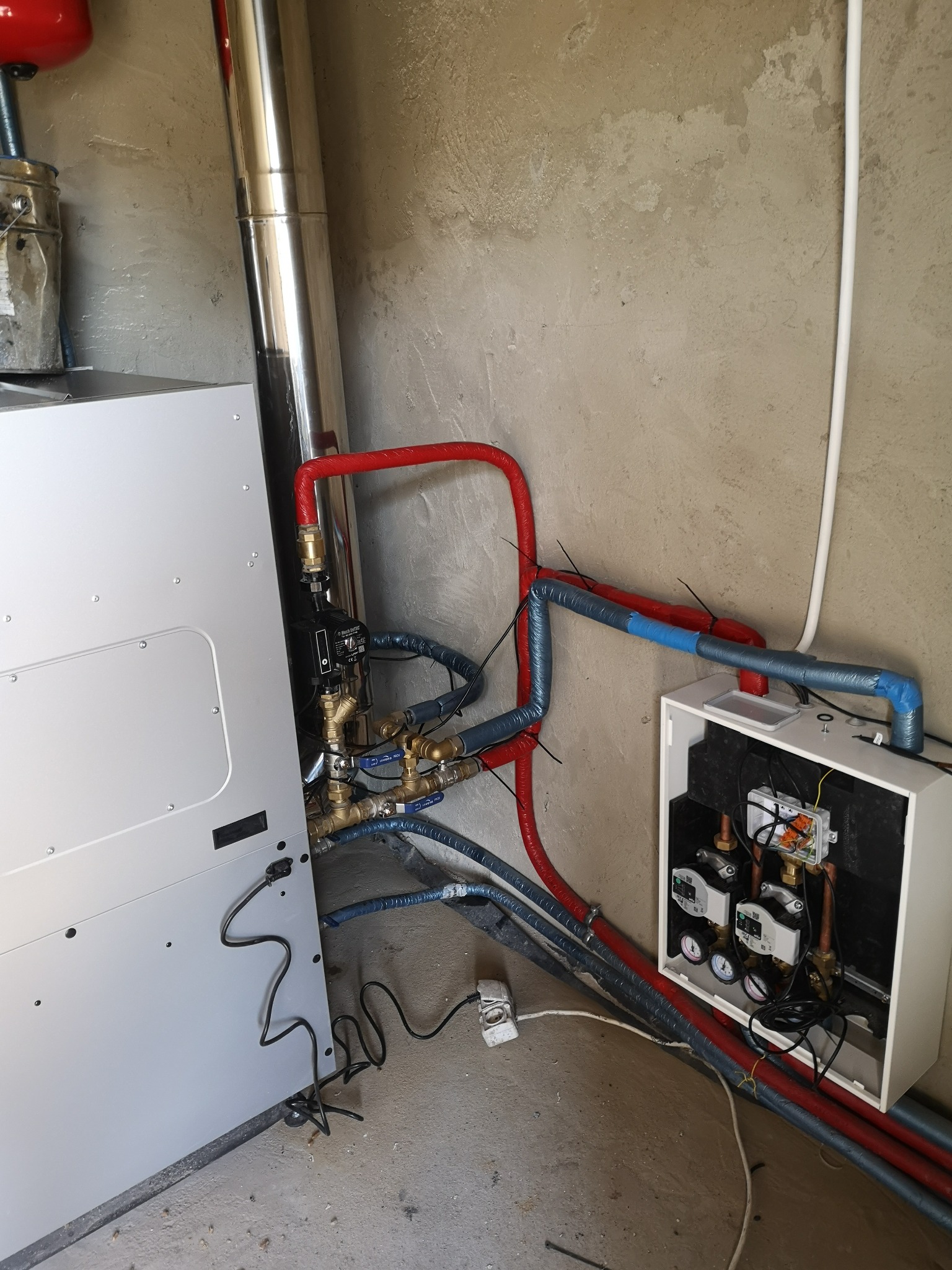 Szczegółowe ujęcie kotłowni z widocznymi rurami w izolacji czerwonej i niebieskiej, zaworami, manometrami oraz białym panelem sterującym, na tle surowej, betonowej ściany.