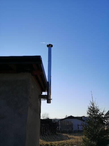 Zewnętrzny, metalowy komin zamontowany na ścianie budynku z widocznymi elementami mocującymi, na tle błękitnego nieba i zarysu zabudowań w oddali.