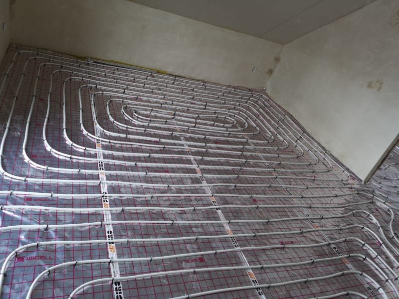 Rozłożona instalacja ogrzewania podłogowego z białych rur na folii z siatką w surowym pomieszczeniu przed wylewką.