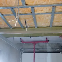 Montaż płyty gipsowo-kartonowej na metalowym stelażu sufitowym z widoczną izolacją z wełny mineralnej, podtrzymywana przez czerwony podnośnik, widoczne kable elektryczne.