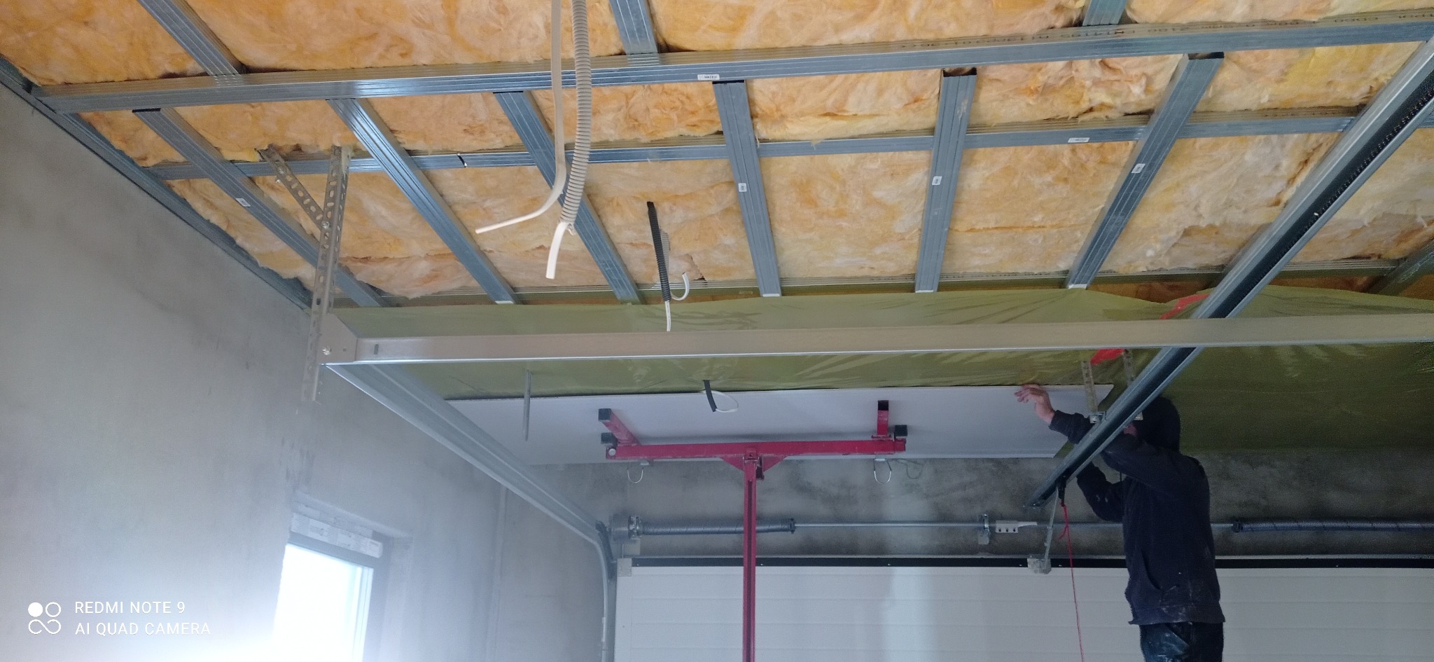 Montaż płyty gipsowo-kartonowej na metalowym stelażu sufitowym z widoczną izolacją z wełny mineralnej, podtrzymywana przez czerwony podnośnik, widoczne kable elektryczne.