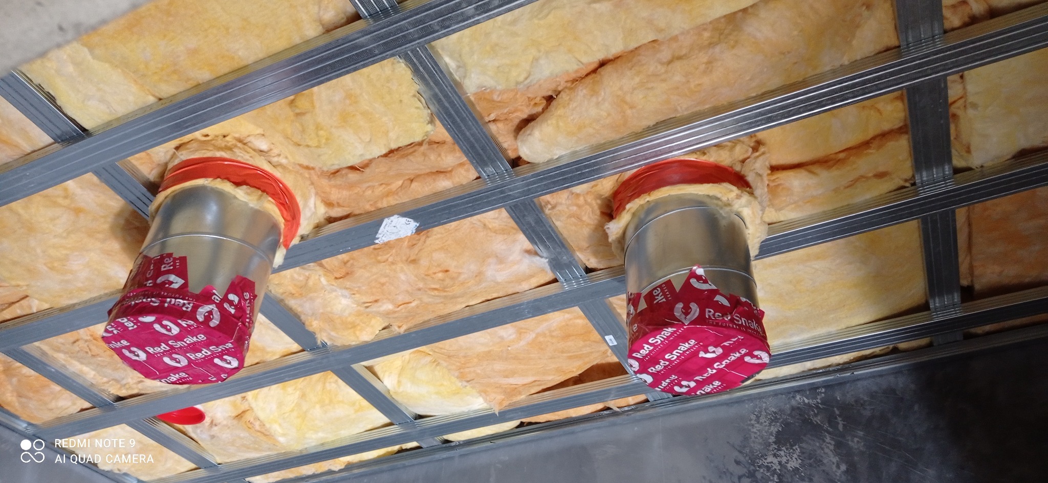 Ocieplenie poddasza wełną mineralną z widocznymi srebrnymi profilami konstrukcyjnymi i izolacją rur wentylacyjnych z czerwonymi etykietami Red Snake.