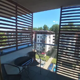 Zadaszony balkon z drewnianymi lamelami, widok na osiedle mieszkaniowe w słoneczny dzień, mały stolik kawowy i dwa krzesła na balkonie.