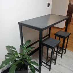 Kompaktowy zestaw barowy z czarną metalową ramą i blatem imitującym kamień, uzupełniony o dwa stołki barowe w tym samym stylu i doniczkę z zieloną rośliną.