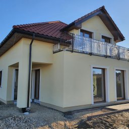 EKO-DOM Kamil Stańczyk - Żółta elewacja domu jednorodzinnego z balkonem, częściowo zabezpieczonym folią, na tle błękitnego nieba i dwóch pracowników w pomarańczowych kombinezonach układających kostkę brukową.