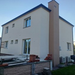 EKO-DOM Kamil Stańczyk - Dwukondygnacyjny dom jednorodzinny po nałożeniu jasnego tynku elewacyjnego, z widocznym kominem w kolorze zbliżonym do elewacji, z przygotowanym miejscem na taras i ułożonymi cegłami.