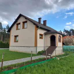 Ocieplony dom jednorodzinny w Lisowie z beżową elewacją, brązowymi oknami i dachem, widok z zewnątrz na ogrodzony ogród i las w tle.