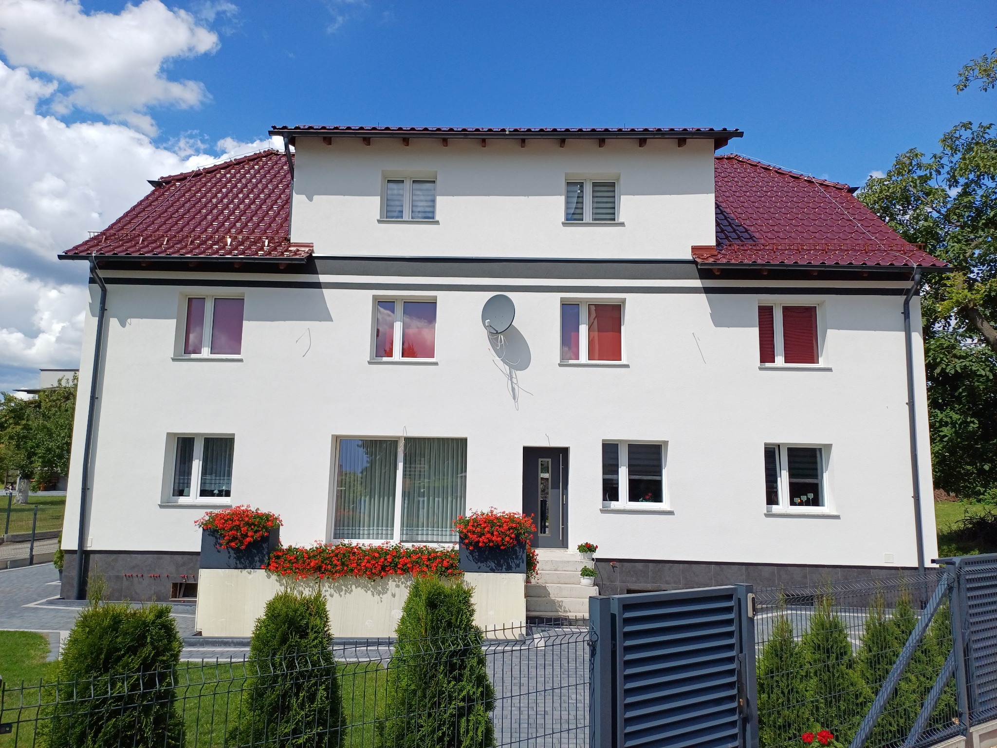 Dwupiętrowy dom z białą elewacją, czerwoną dachówką, kwiatami w donicach i anteną satelitarną na ścianie, Pawonków.