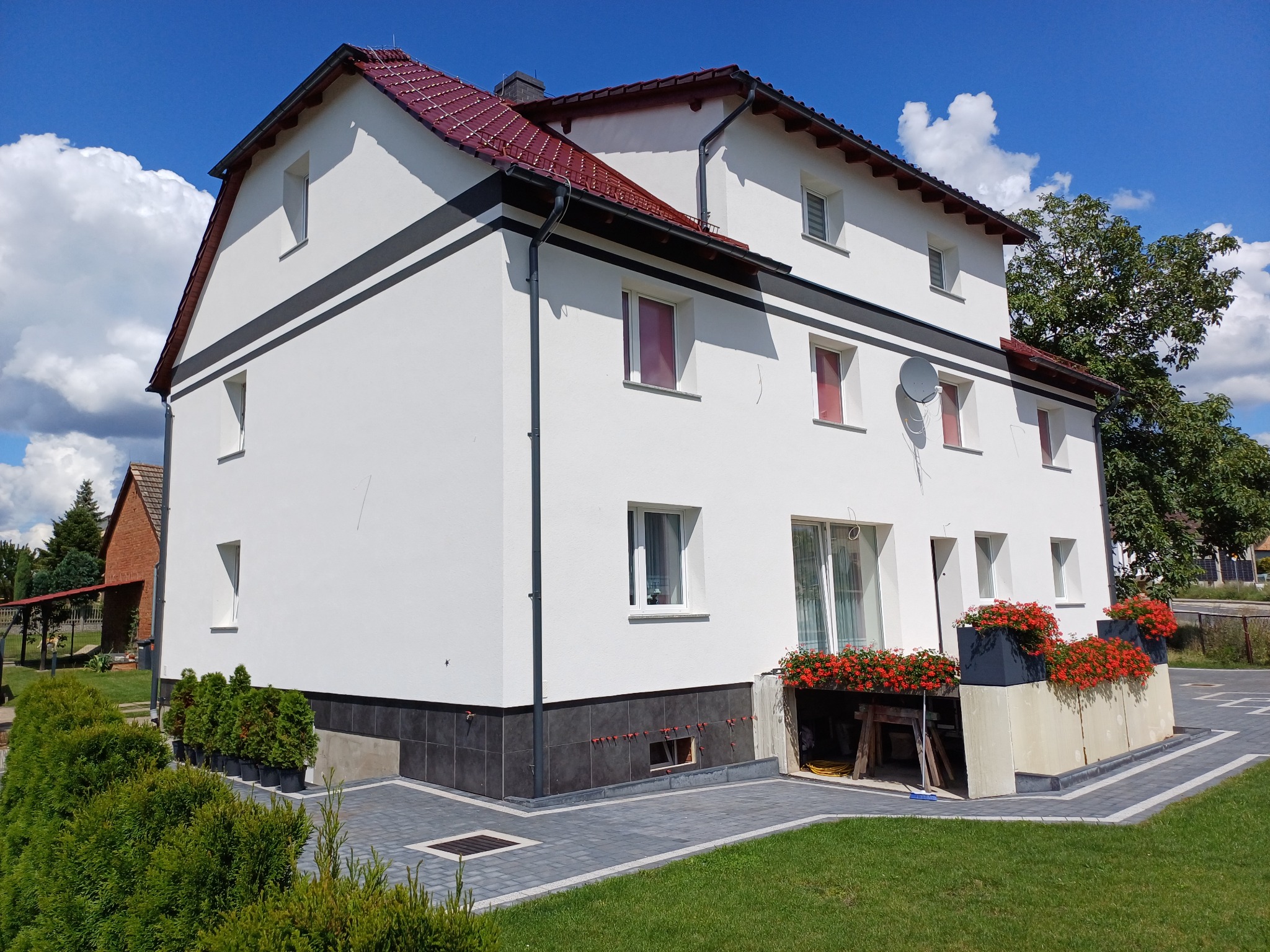 Dwukondygnacyjny dom po remoncie elewacji, z białymi ścianami, ciemnymi pasami i czerwoną dachówką, otoczony zielenią, z widocznymi detalami wykończeniowymi.