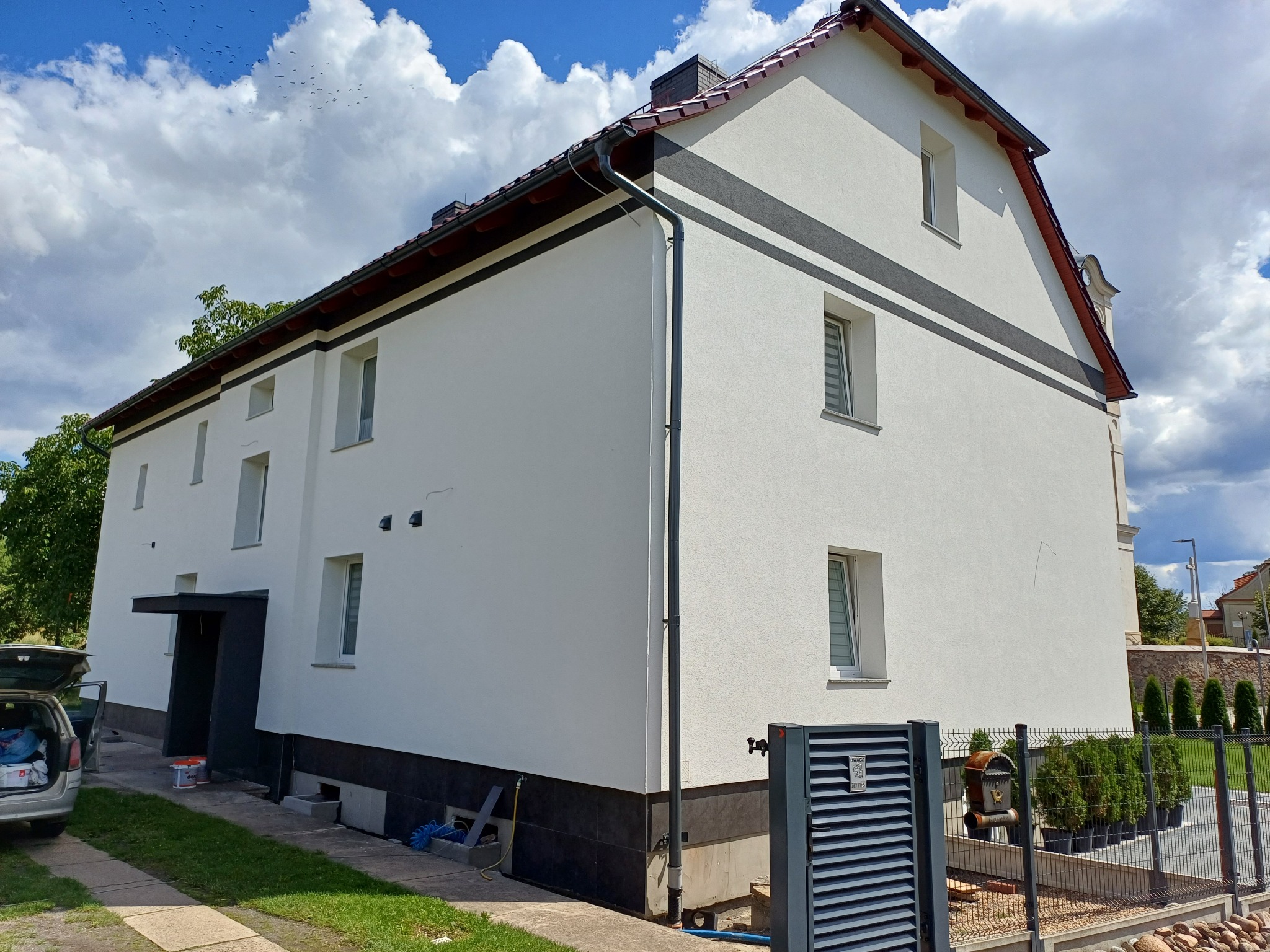 Dwukondygnacyjny budynek po renowacji elewacji, z białymi ścianami, ciemnoszarymi akcentami, brązowym dachem i widocznymi elementami otoczenia, takimi jak trawnik, podjazd i częściowo otwarty...