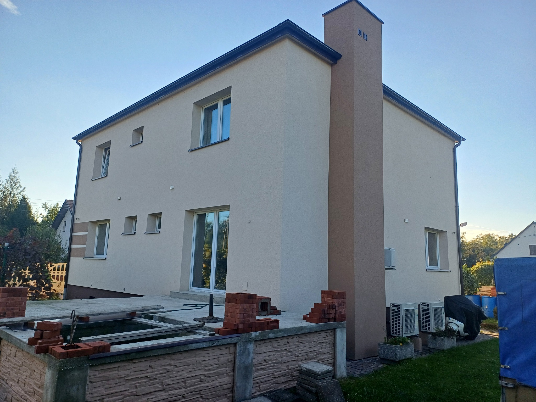 Dwukondygnacyjny dom jednorodzinny po nałożeniu jasnego tynku elewacyjnego, z widocznym kominem w kolorze zbliżonym do elewacji, z przygotowanym miejscem na taras i ułożonymi cegłami.