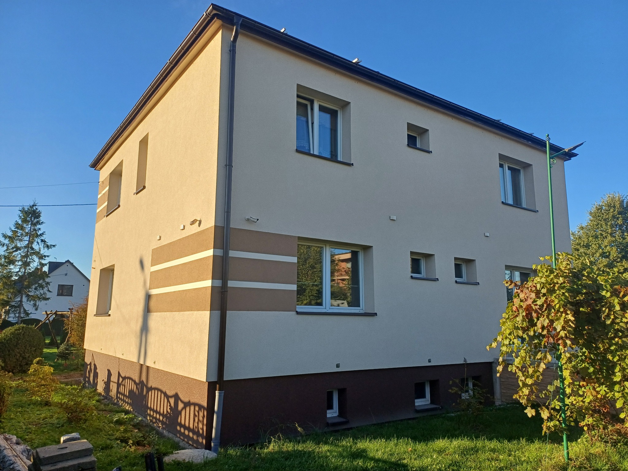 Dwukondygnacyjny dom z jasną elewacją, brązowym pasem u dołu i trzema poziomymi pasami w odcieniach brązu, widoczny w słoneczny dzień, z ogrodem w pierwszym planie.