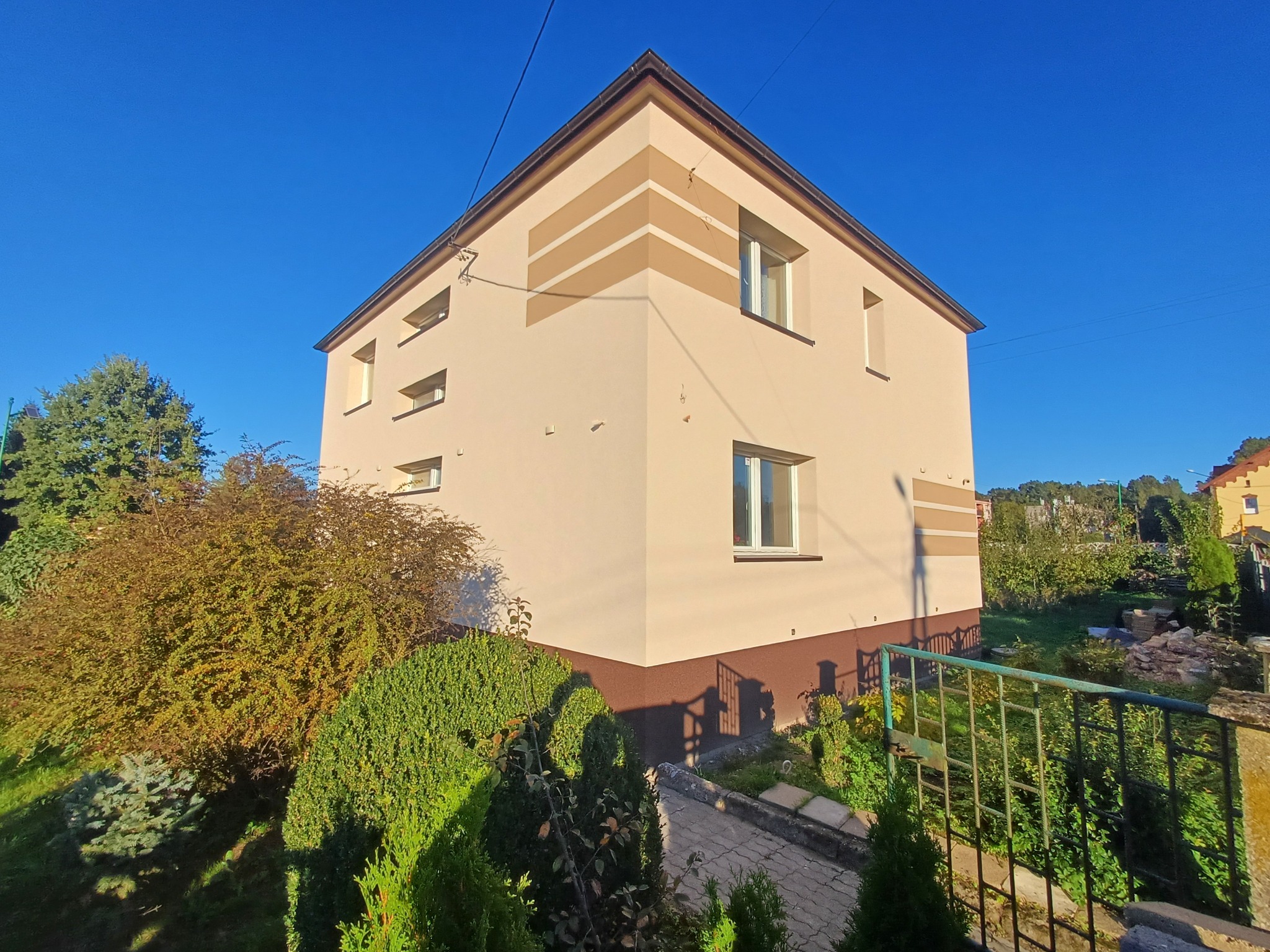 Dwukondygnacyjny dom z beżową elewacją i poziomymi, brązowymi pasami dekoracyjnymi, widok z poziomu ogrodu przy metalowym ogrodzeniu, błękitne niebo w tle.