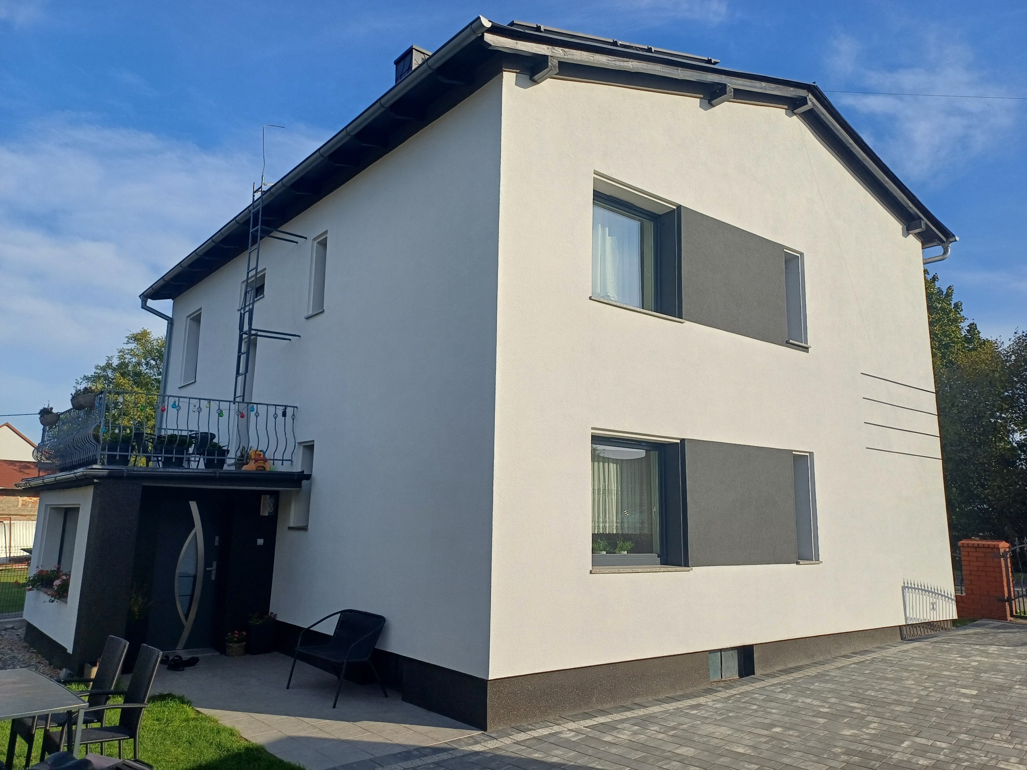 Dwukondygnacyjny dom po renowacji elewacji, z jasną elewacją i ciemnymi akcentami wokół okien i dachu, z drabinką przymocowaną do ściany bocznej i balkonem nad wejściem.