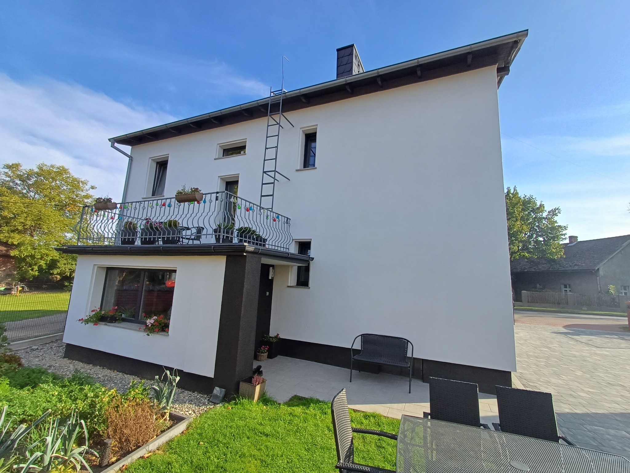 Dwupiętrowy dom z białą elewacją, czarnymi akcentami, balkonem z kwiatami i drabiną na dachu, otoczony zielonym ogrodem, w słoneczny dzień.
